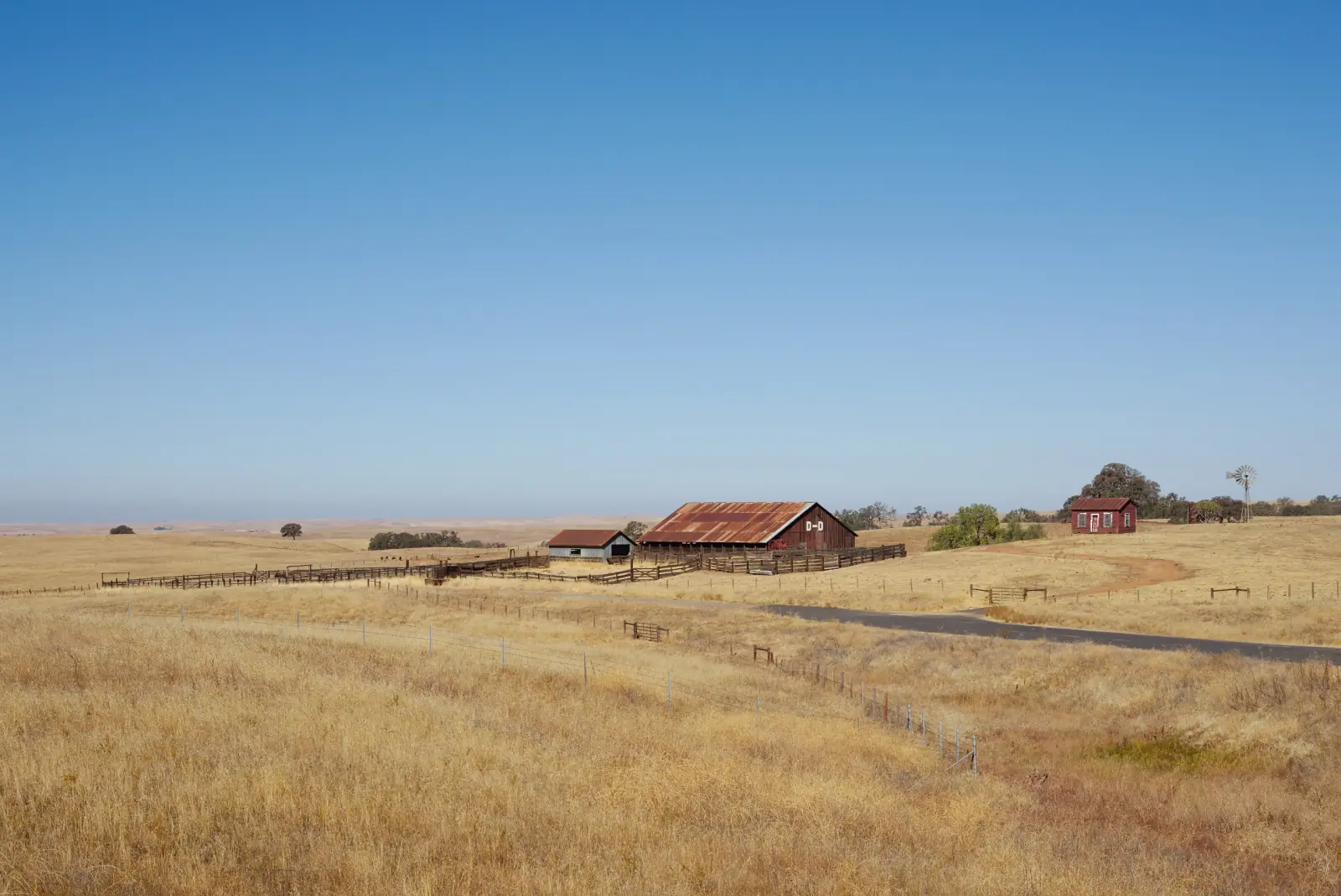 A rust-roofed barn and corrals stand on golden grassland under a clear blue sky in the foothills of Calaveras County.