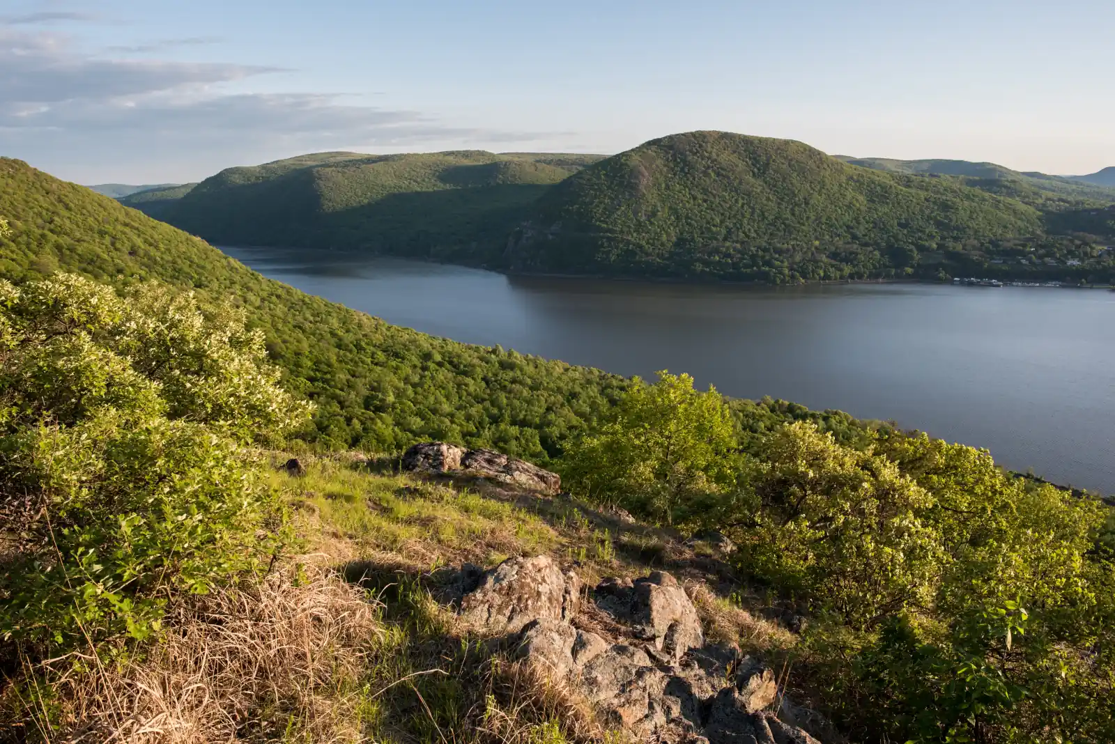 Sunlit forested hills of the Hudson Highlands rising above the Hudson River, with Storm King Mountain across the water, seen from Sugarloaf Mountain in Dutchess County, New York.