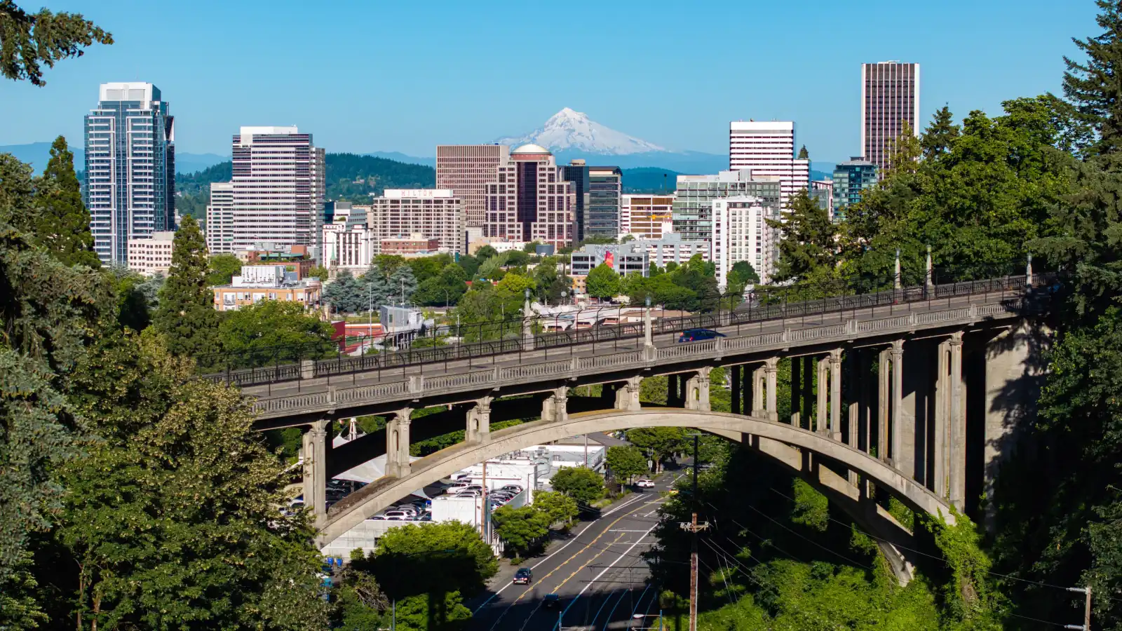 Aerial view of Portland's Vista Bridge with downtown towers and snow-capped Mount Hood behind it under a clear blue sky.