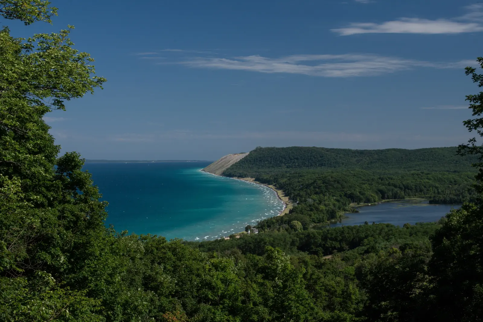 Aerial-style view from Empire Bluffs over turquoise Lake Michigan, the Sleeping Bear dune bluff and forested hills on the Leelanau Peninsula.