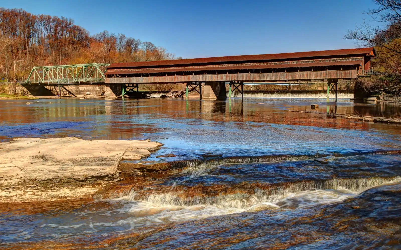 Harpersfield Covered Bridge spanning the Grand River in autumn, with rock ledges and a small waterfall in the foreground.